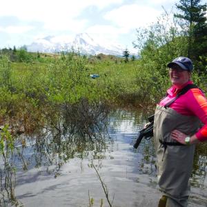 Angela wearing waders and a red shirt with blue cap in the middle of a stream.