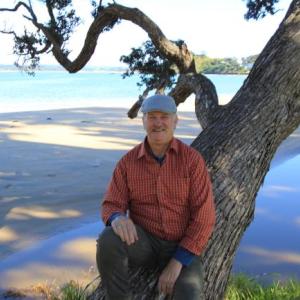 David leaning against a tree with a body of water and sandy shoreline behind him.