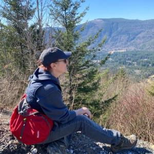 Dawna sitting on a rock overlooking a valley wearing a blue coat, hat and red backpack