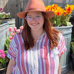 Melissa wearing a orange hat and a pink and purple and white stripped shirt standing outside in front of containers of colorful tulips